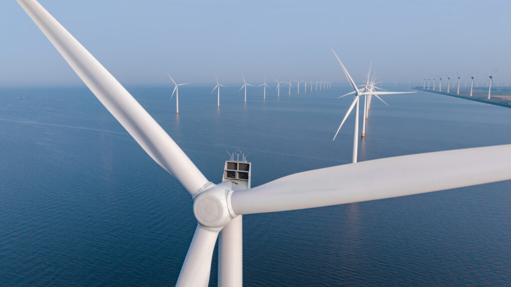 Close-up view of a wind turbine blade with an offshore wind farm stretching across calm blue sea.