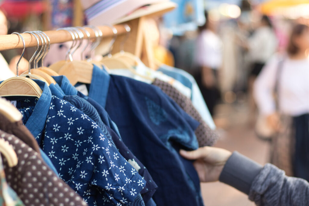 Clothes hanging on wooden racks at an outdoor market with blurred shoppers in the background, blue patterned fabrics visible in foreground.
