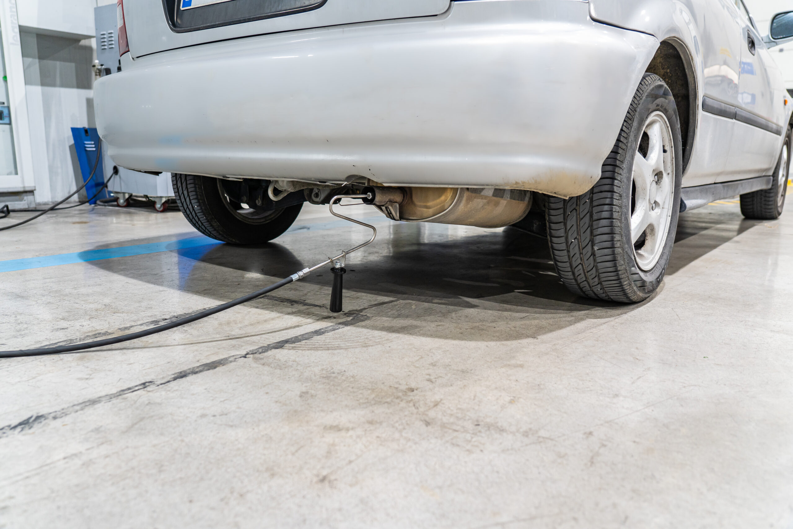 Rear view of a silver car on a workshop floor with exposed exhaust and a support stand beneath the bumper.