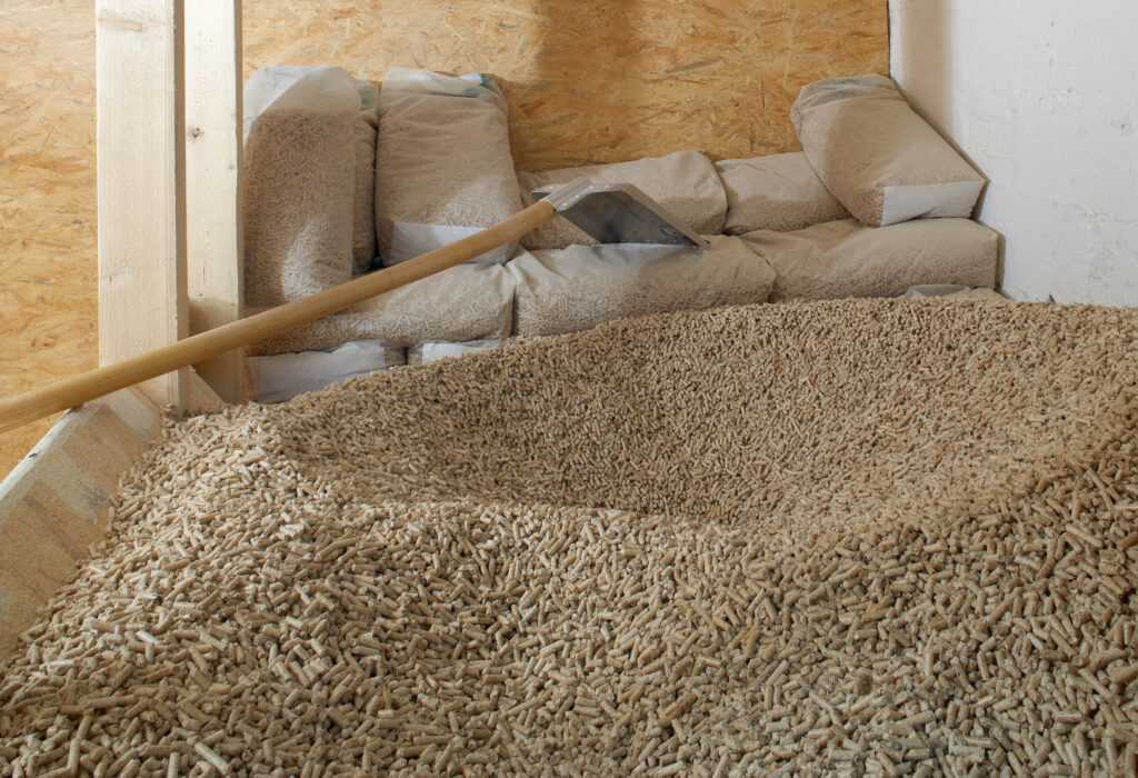 Indoor wood-pellet storage: loose pellets covering the floor with bagged pellets stacked along the back wall and a shovel resting on the piles