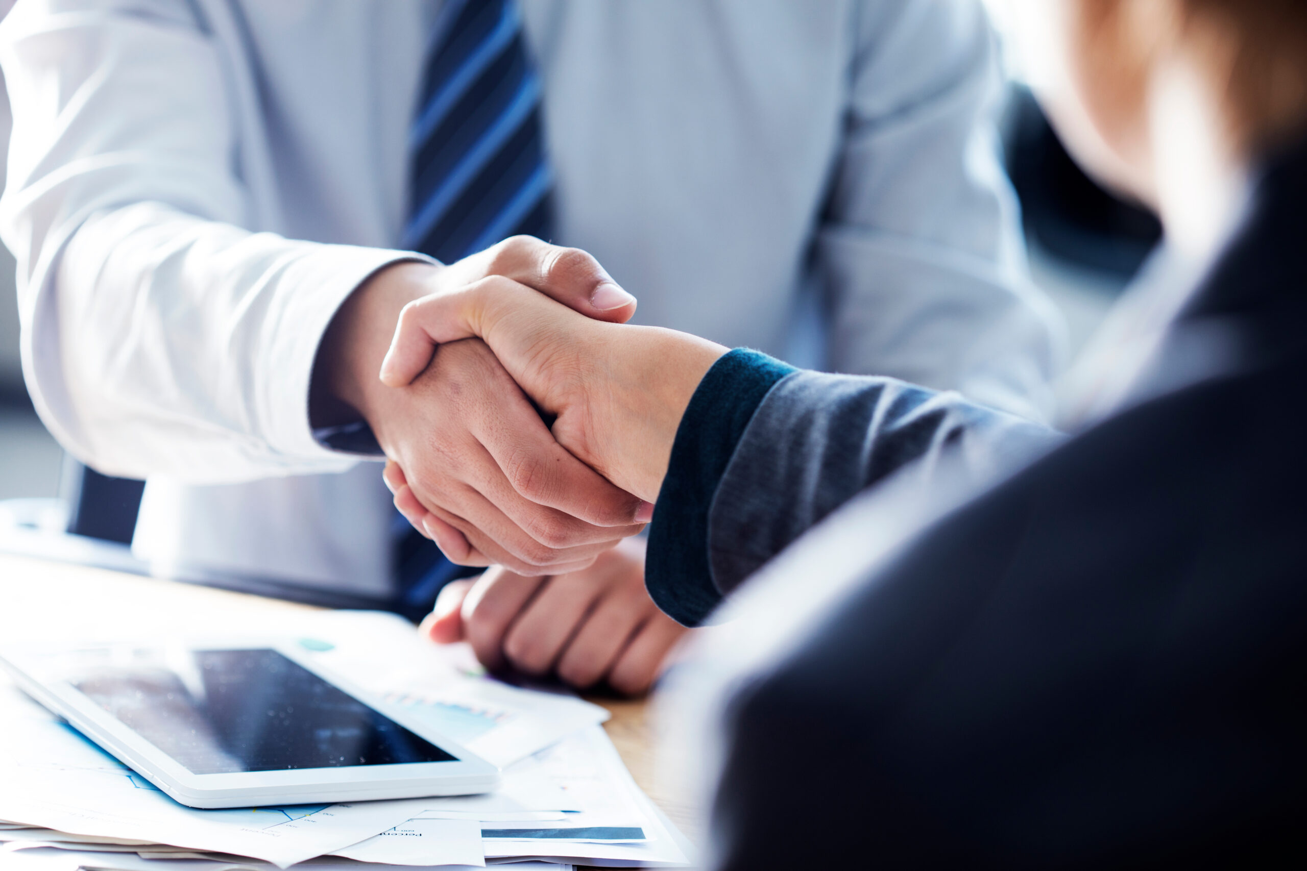 Two professionals shake hands over a desk with papers and a tablet, signaling an agreement during a business meeting.