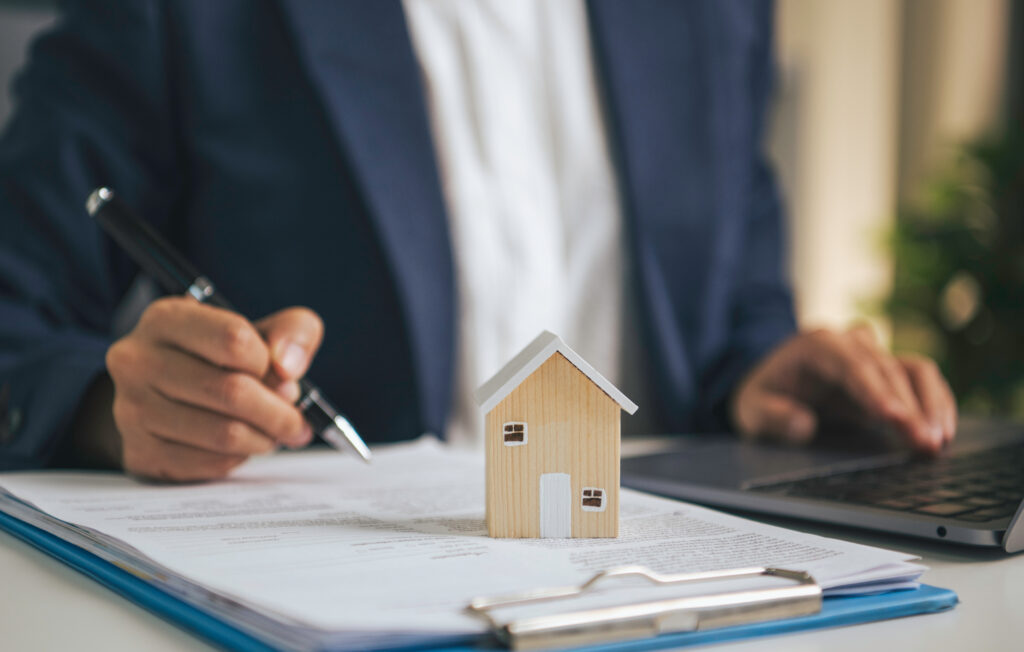 Person in a navy suit signs documents with a pen beside a small wooden house model on a desk.