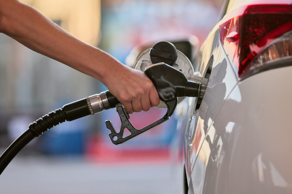 Person's hand holding a fuel nozzle as it pumps gas into a car's fuel tank at a station.