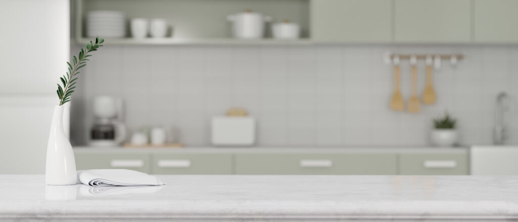 White vase with a green sprig on a marble kitchen island, with a blurred pastel-green kitchen in the background.