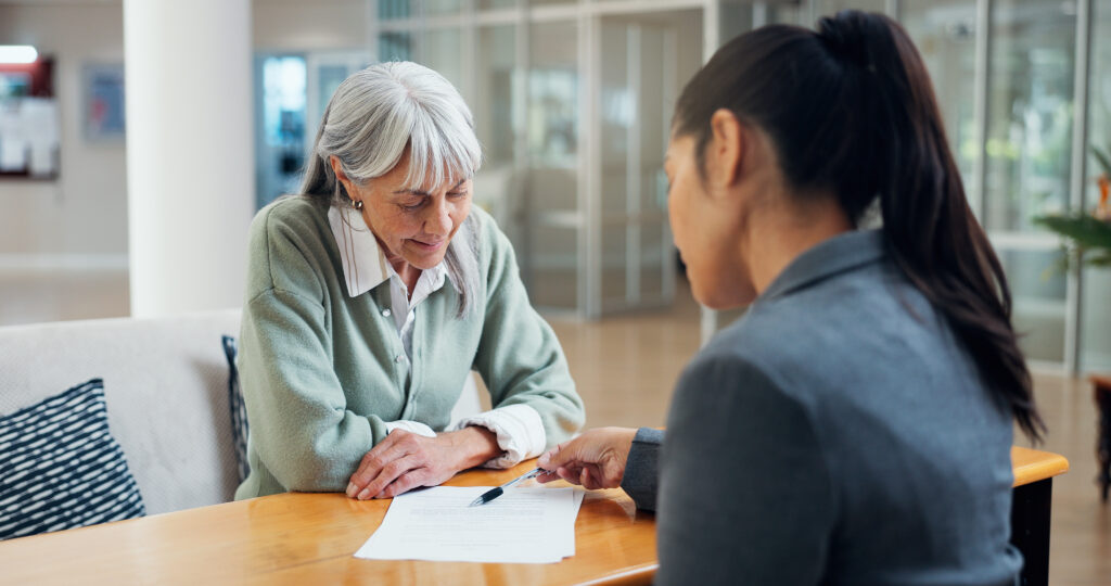Elderly woman with gray hair and green cardigan sits at a table, reviewing documents with a practitioner across from her in a bright lobby.”,