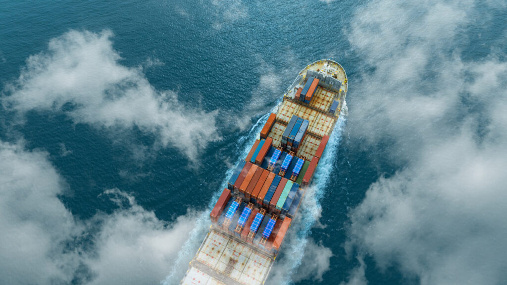 Aerial view of a multicolored container ship cutting through blue ocean, with stacks of shipping containers on deck under scattered clouds.