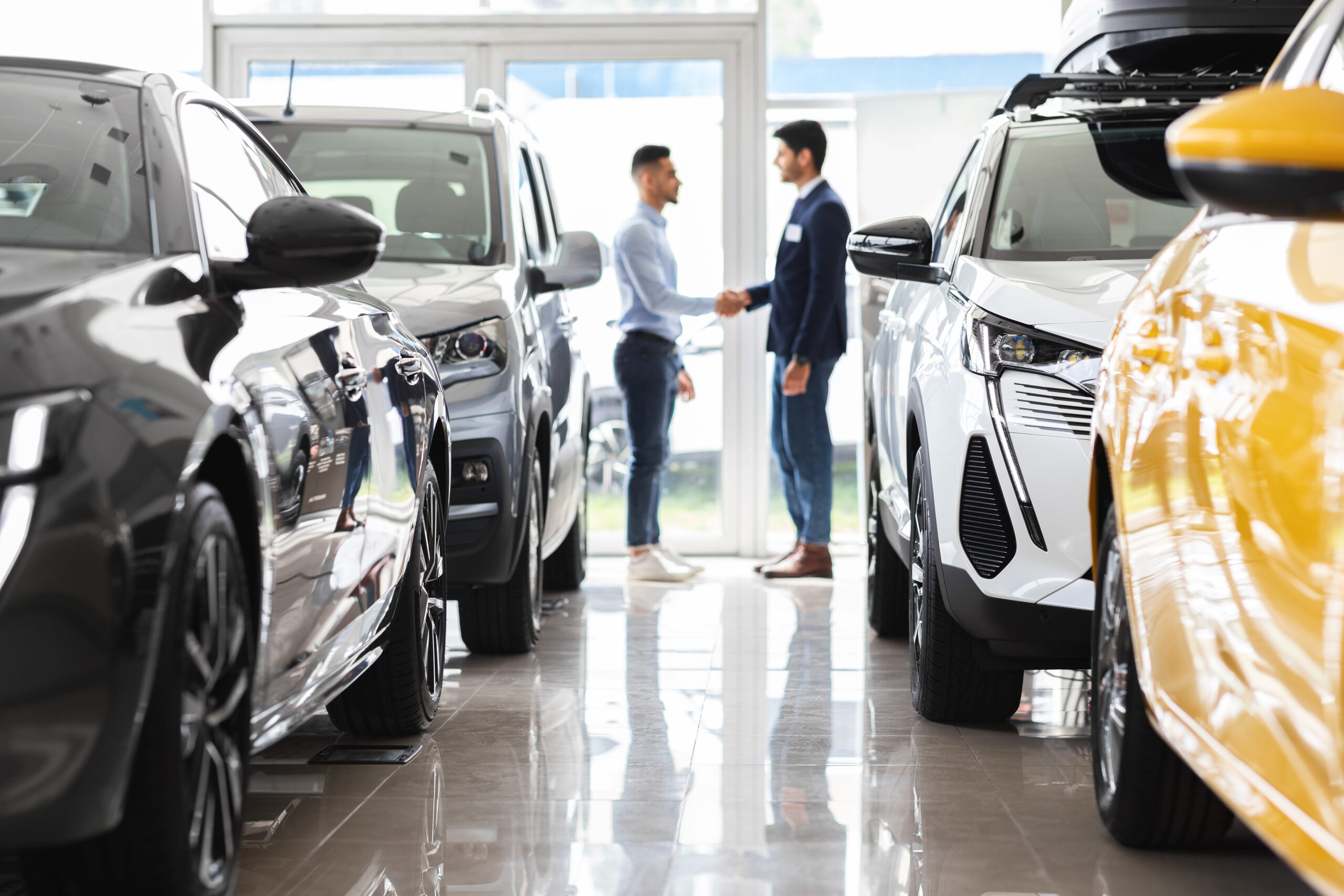 Two men shake hands in a car showroom between rows of new SUVs and cross-aisle light reflections.