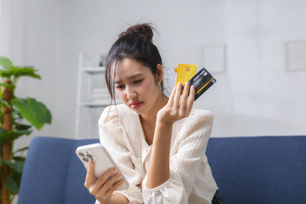 Woman seated on a blue couch, looking at her smartphone while holding several credit cards in her right hand.