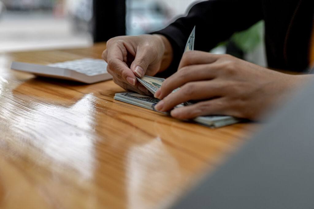 Hands counting multiple US dollar bills on a wooden table with a keyboard in the background.
