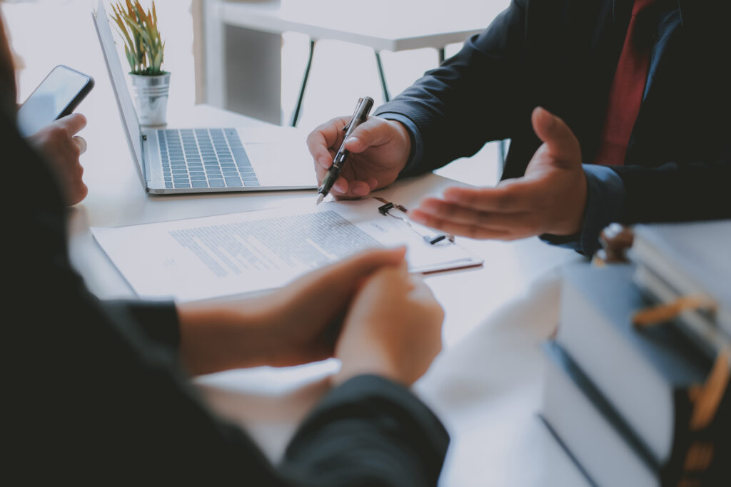 Business colleagues signing documents at a table with a laptop nearby and a clipboard in view, during a meeting.