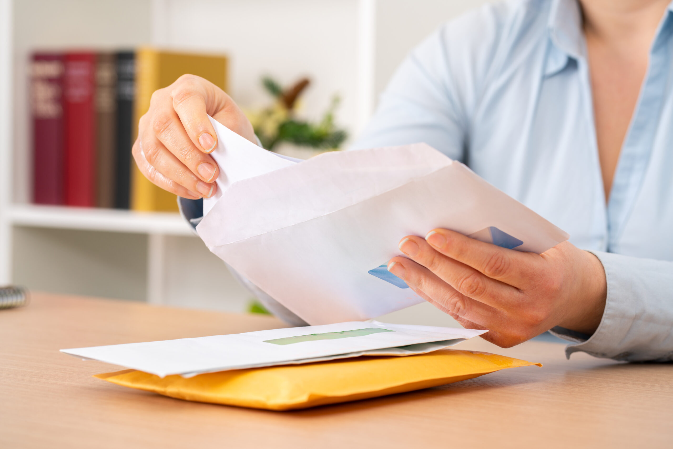 Person tearing open a white envelope at a desk, with more envelopes nearby on the desk and a blurred bookshelf in the background.