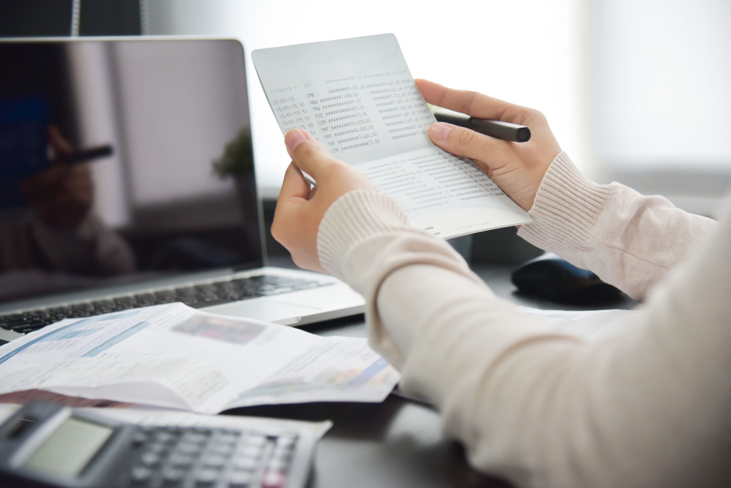 Hands hold a printed financial report with rows of numbers, pen ready on a desk near a laptop.