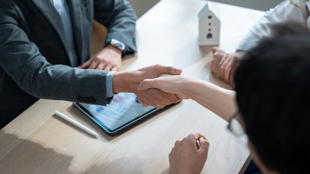 Two people shake hands over a wooden table, with a tablet and stylus nearby, suggesting a business agreement or meeting.