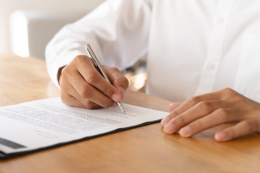 Person signing a contract with a silver pen on a wooden desk.