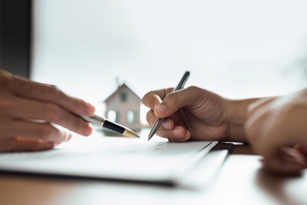 Two hands signing documents with pens on a desk, a small house model in the blurred background.