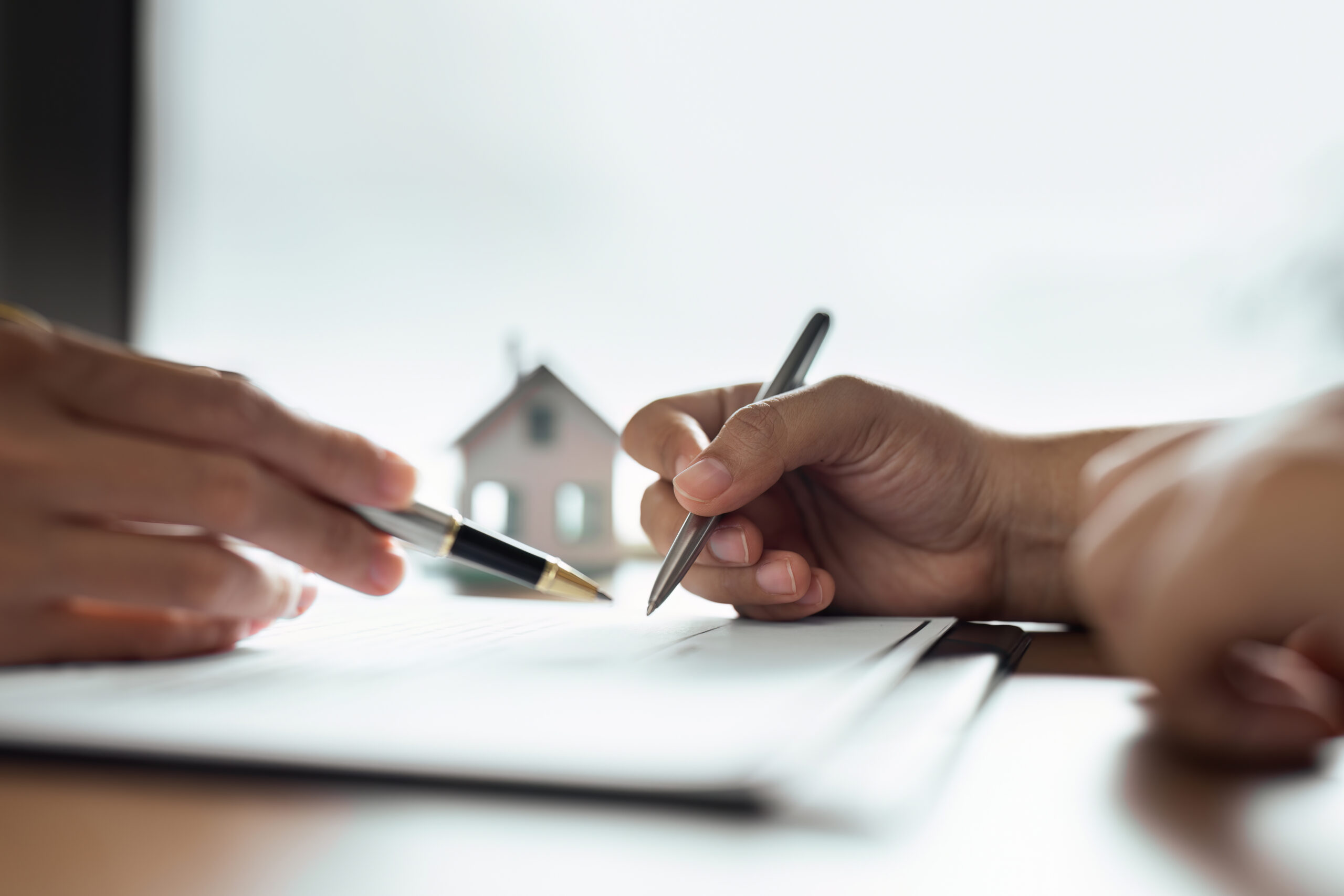 Two hands signing documents with pens on a desk, a small house model in the blurred background.