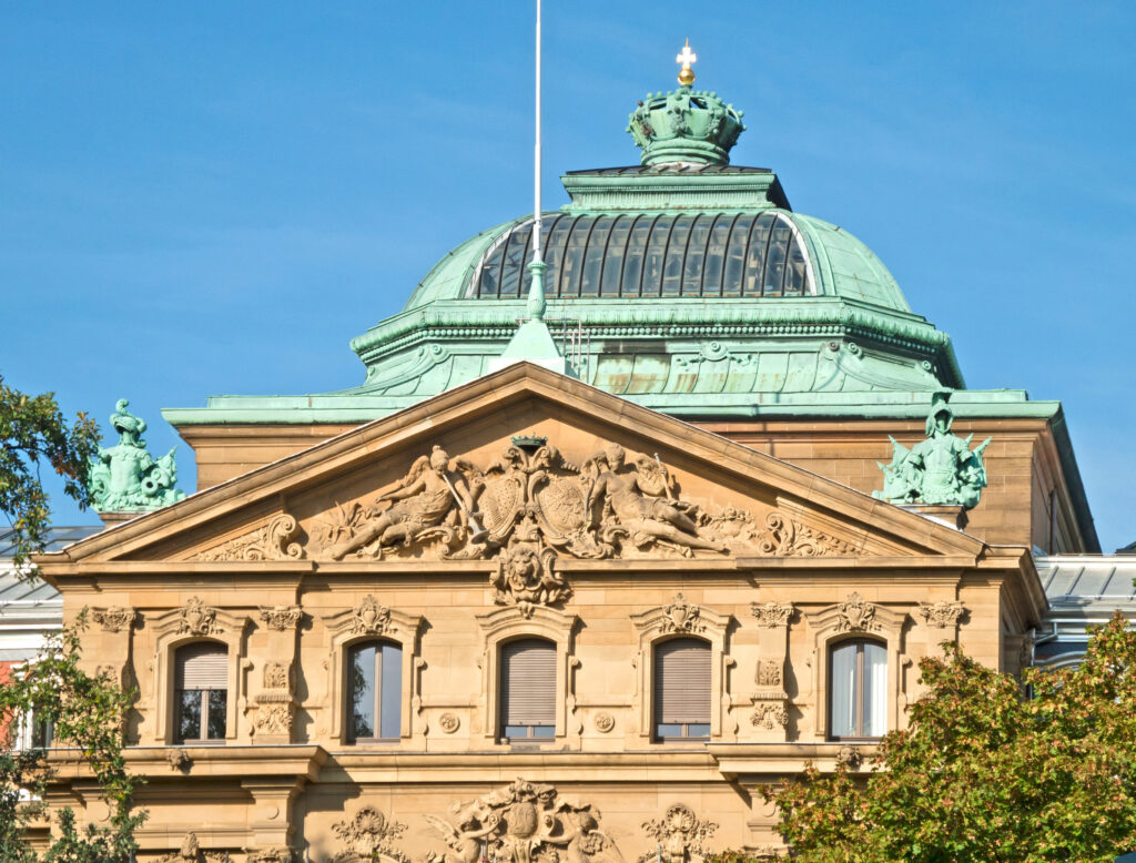 Front façade of a historic stone building with a green copper dome and sculpted reliefs under a clear blue sky.