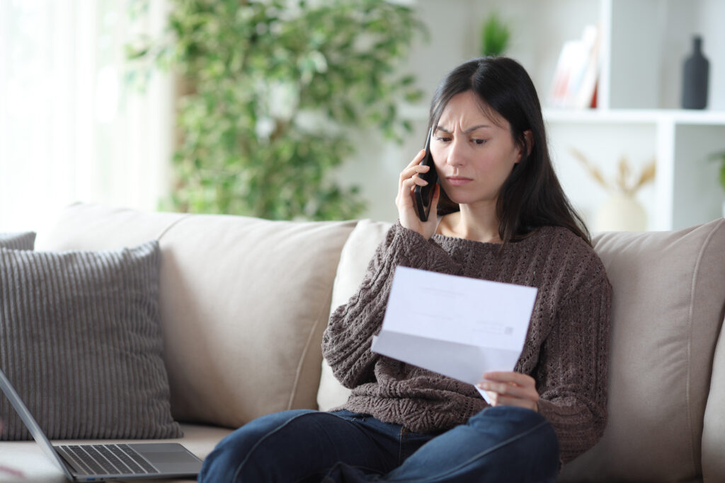 Woman sitting on a beige sofa, talking on her cellphone while holding a letter and a laptop nearby.