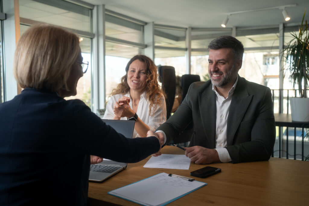 Three professionals in a modern office shake hands across a wooden table, with a laptop, papers, and a smartphone nearby.