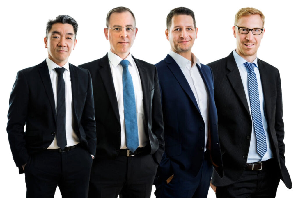 Four businessmen in suits standing side by side, smiling slightly for a corporate photo on a white background