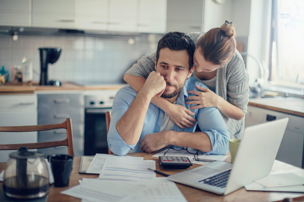 Man at a kitchen table looks stressed while a partner hugs him from behind, with papers, a calculator, and a laptop in view.