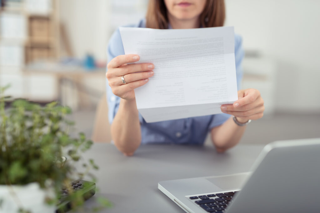 Person holding a printed document toward the camera at a desk with a laptop nearby in an office setting, blurred background.