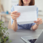 Person holding a printed document toward the camera at a desk with a laptop nearby in an office setting, blurred background.