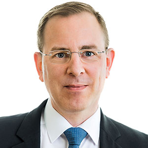 Head-and-shoulders portrait of a man in a dark suit and blue tie, wearing glasses, with a neutral white background.