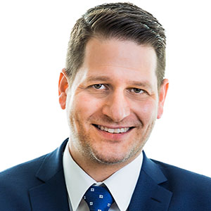 Head-and-shoulders portrait of a smiling man in a navy suit and blue tie against a white background.