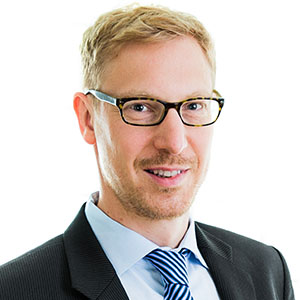 Professional headshot of a man with light hair and glasses, wearing a dark suit and blue striped tie, smiling at the camera.